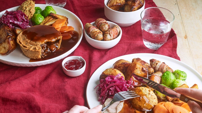 A person tucks into a Christmas lunch filled with turkey, roast potatoes and all the trimmings. On the table is a vegan pie dinner, and a side of pigs in blankets.